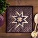 The beautiful Folded Star Squared Hot Pad made with Plumberry fabrics on a wood table