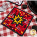 The red and yellow Peace, Love & BBQ Folded Star Squared Hot Pad on a red and white checkered table
