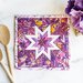 Purple and pink hot pad on a white marble countertop with wooden spoons and purple flowers