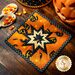 Orange Halloween themed hot pad with central folded star on wood table by jack-o-lantern, spoons, and candy corn.