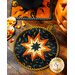 Black and orange Halloween themed hot pad on wood table next to spoons, candy corn, and a jack-o-lantern.