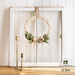 Beige wreath made with a braided design and green leaves made of fabric accented with a beige ribbon hanging from a frame.