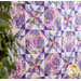 A purple, white, and green quilt hanging on a wall at an angle from the viewer with green leaves out of focus in the foreground