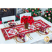 Christmas themed table runner featuring bell designs made of red and cream snowflake fabrics on table with flowers and matching napkin place settings.