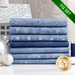 Stack of 7 folded light blue fabrics with metallic silver snowflakes, script, and stars/burst patterns with spools of blue and silver thread next to it with silver baubles on a white countertop with a pale decor in the background and a green banner in one corner that reads: 