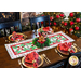 Christmas themed table runner on a brown table with four place settings all around with red napkins, a wreath and gingerbread man in the center, and black chairs and a fireplace in the background