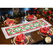 Christmas themed table runner on a brown table with four place settings all around with red napkins and black chairs and a fireplace in the background