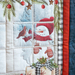 A quilted panel featuring a snowman in a red hat with a bird, surrounded by pine branches and ornaments.