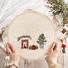 A circular hand embroidery project with the word Joy and a small fireplace with Christmas presents