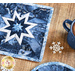Square blue hot pad with a white star in the center on a brown table with a cup of hot cocoa and snowflake decorations.