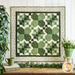Small quilt wall hanging featuring pieced shamrocks with geometric designs all in green and cream. A potted plant, green house tree, and a green mug atop a small stack of fabric from the Lucky Charms 