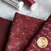 Close up of dark red fabric with metallic silver winter motifs layered atop one another on a white countertop with red and silver spools of thread and a mesh silver ribbon laying next to them