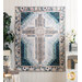 isolated image of a rectangular quilt that features a large cross motif in the center in muted floral fabrics with dark teal accents hanging on a white paneled wall with home decor items on a shelf and small chair with a house plant on either side of the quilt