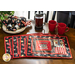 image of four placemats made with the Coffee Always fabric collection featuring red, black, and neutral tones with coffee mugs and coffee beans, fanned out on a brown table with ornate cream plates, napkins, red mugs, and a french press with a green plant and window in the background