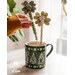 two decorative shamrocks with leaves made of felt and a fabric covered dowels held by a hand placing them inside a green mug decorated with gnomes on a white table in front of a window and green house plant.