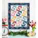 Blue, white, yellow, and red mini quilt with blocks featuring a swirling pattern with different fabrics in each one hanging on a white paneled wall above a white countertop with fresh garland, a scale with pink flowers, and a vase of red tulips on either side.