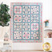 A bright, white paneled wall with a patchwork quilt with classic quilt star blocks in soft blue, pink, and cream hanging flat with a small white shelf, white chair, white blanket, and decor in soft pink and blue shades that match the quilt with a dark green houseplant on the left