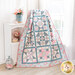 A bright, white paneled wall with the completed Pretty Patchwork quilt draped over furniture with a small white shelf and flowers with decor in soft pink and blue shades that match the quilt