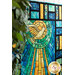 45 degree side image of the peaceful window quilt, made to look like stained glass from blue, aqua, and yellow piecing, all bordering a yellow dove with sunray designs behind it. The quilt is hanging from a wall and framed by a tree on the left