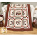 Photo of a square quilt featuring a house and floral motifs in each block draped over a white bench with floral burgundy and white decor all around