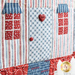 Close up of detail on the front door of the quilted house showing two small buttons, one heart shaped, on a blue patterned door with red windows and red, white, and blue striped walls