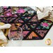 close up of one end of the table runner showing intersecting black fabric strips and brightly colored, stylized flowers in triangular sections with a decorative glass and gold rimmed tables with cream napkins on a white table