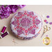 A round purple and pink pincushion quilted in an intricate flower shape on a beige countertop with purple themed notions scattered nearby and purple flowers in the top left corner