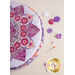 A round purple and pink pincushion quilted in an intricate flower shape just off camera on a beige countertop with purple themed notions scattered nearby