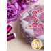 A round purple and pink pincushion quilted in an intricate flower shape just off camera on a beige countertop with purple spools of thread and purple flowers in the background