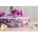 Side view shot of a round pink and purple pincushion showing an intricate floral fabric on a beige countertop with scissors and buttons on the counter, purple flowers in the bakground, and pins stuck in the top of the pincushion