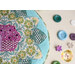 A round green and teal pincushion quilted in an intricate flower shape just off camera on a beige countertop with multicolored flower shaped and round buttons in the background