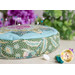 Side view shot of a round green and teal pincushion showing an intricate floral fabric on a beige countertop with colored buttons on the counter, purple flowers and other greenery in the background, and pins stuck in the top of the pincushion