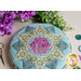 A round green and teal pincushion quilted in an intricate flower shape with purple accent colors on a beige countertop with green, blue and purple buttons scattered nearby with gold scissors and purple flowers in the top left corner