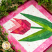 Finished quilt block featuring a pink tulip with green leaves bordered by pink mottled fabrics laying on a wooden countertop and framed by greenery