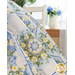 Close up of a blue, cream, and green floral quilt draped out of frame with a white chair, pitcher, and blue flowers in front of a window in the background