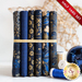 6 folded dark blue fabrics with gold metallic motif accents propped vertically bound with a gold ribbon on a white countertop with gold and blue spools of thread and a large blue bauble to the right of it, with a gold star decoration in the background and a red banner in the top right corner that reads 