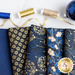 A close up of dark blue and gold metallic fabric with winter motifs fanned out with spools of gold and blue thread and a large blue bauble in the background.