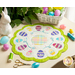 A round scalloped table topper with decorated easter eggs on a white background with a bright green border and hand embroidered details on a cream countertop with scattered notions and easter eggs all over with a ceramic rabbit and green houseplant with an easter basket in the top right corner.
