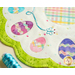 Close up of table topper edge showing a bright green border and decorated easter eggs on a cream background with tiny multicolored bunnies all over and a plaid round center on a cream countertop with a spool of blue embroidery thread.