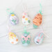 Six Easter Egg Ornaments in different styles and pastel colors laying on a white marble countertop