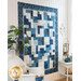 Hanging quilt filled with asymmetrical blocks in blue and cream batiks against a white paneled wall with a white wicker chair, tall houseplant, and small wicker table with seaside decor on either side.