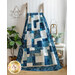 Hanging quilt filled with asymmetrical blocks in blue and cream batiks draped on a wooden ladder against a white paneled wall with a white wicker chair, tall houseplant, and small wicker table with seaside decor on either side.