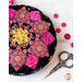 A round pink, yellow and black pincushion quilted in an intricate flower shape on a white marble countertop with pink buttons and ornate scissors scattered nearby and pink flowers in the top left corner