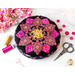 A round pink, yellow and black pincushion quilted in an intricate flower shape on a white marble countertop with pink and yellow themed notions scattered nearby and pink flowers in the top left corner