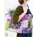 Photo of a person in profile facing to the right with flowers in the back of a purple and green tote bag made with floral fabrics. A white wall is out of focus in the background.