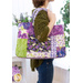 Photo of a person in profile facing to the right with flowers in the back of a purple and green tote bag made with floral fabrics. A white shelf and walls with a houseplant out of focus in the background.
