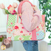 Photo of a person in profile facing to the right with tulips in the back of a pink, green and blue tote bag made with floral, gingham and paisley fabrics. A white shelf and walls with a houseplant out of focus in the background.