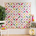 Image of a full white quilt with circular pieced designs, made up of green, pink, and yellow floral patterns. The quilt is hung on a wall, framed on the left by a small tree and a white chair with lemons, and on the right with a white end table supporting a pink watering can with flowers in it