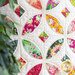 Close up image of the local honey orange blossom throw quilt, consisting of a white background with 5 overlapping circular designs made up of petal and diamond shapes, each made from different floral fabrics in pink, yellow, and green. The image is framed on the left by green leaves out of focus in the foreground 