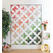 Full size quilt with an ombre of pink and green across rows of cross shapes on a white background hanging on a white paneled wall with a white shelf with home decor and flowers, and a houseplant with a small basket of flowers at the base.